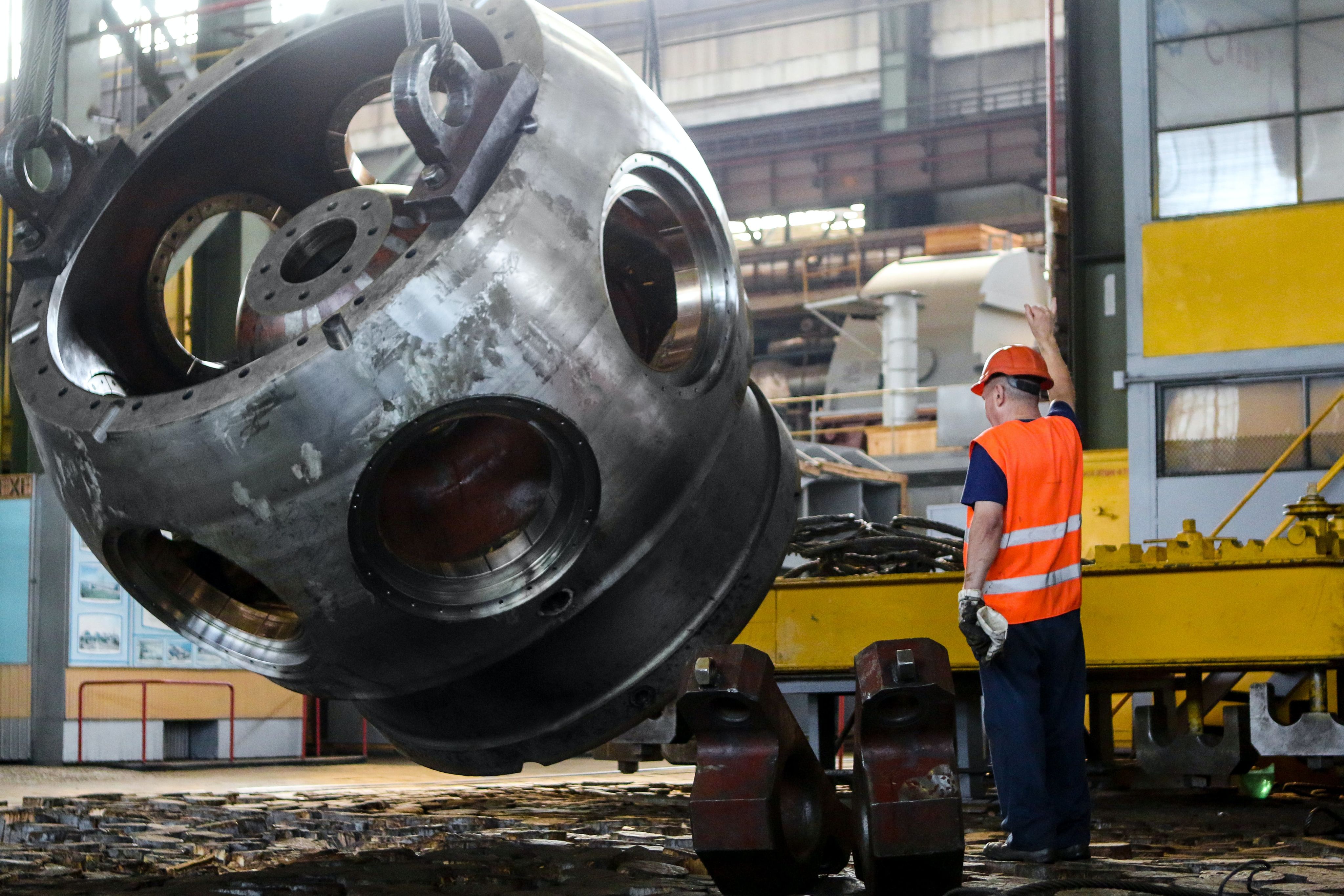 A man in safety gear directs as a crane lifts large steel machinery component.